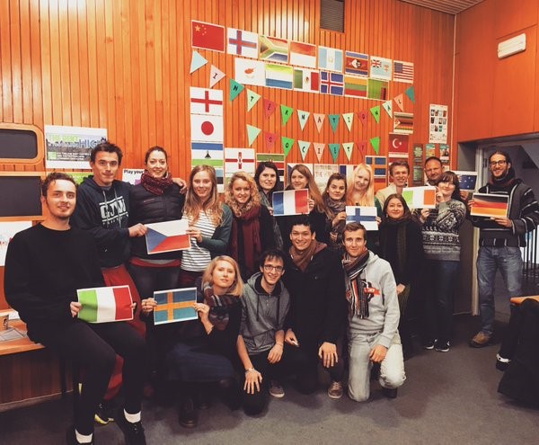 A large group of people in front of a wood panelled wall covered in flags of different nations.