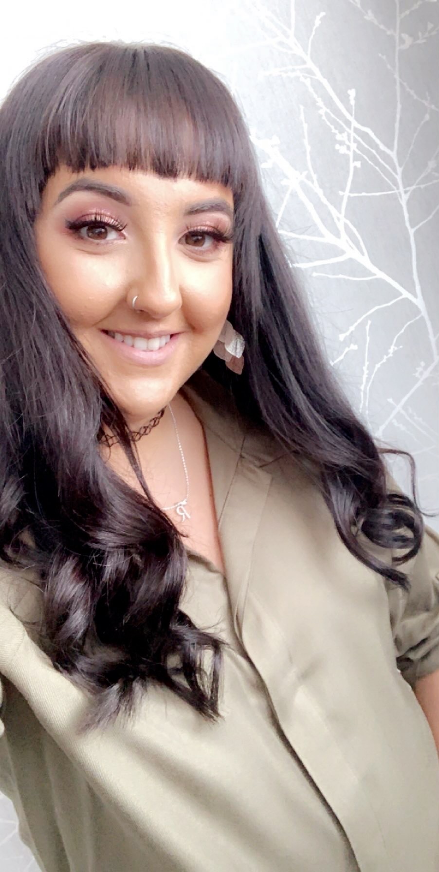An head and shoulders image of Roz looking into the camera and smiling. Roz is a woman with long wavy brown hair with a fringe. She is leaning against a grey wall, wearing a light brown jacket.