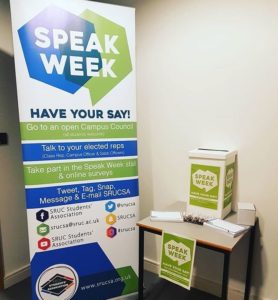 Speak Week stand on campus, there is a small table with a pile of cards on and a box to put them in, beside that is a large sign saying 'speak week'